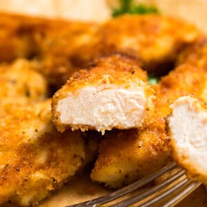 A square image of a chicken cutlet cut in half revealing the cooked inside positioned on top of other chicken cutlets.