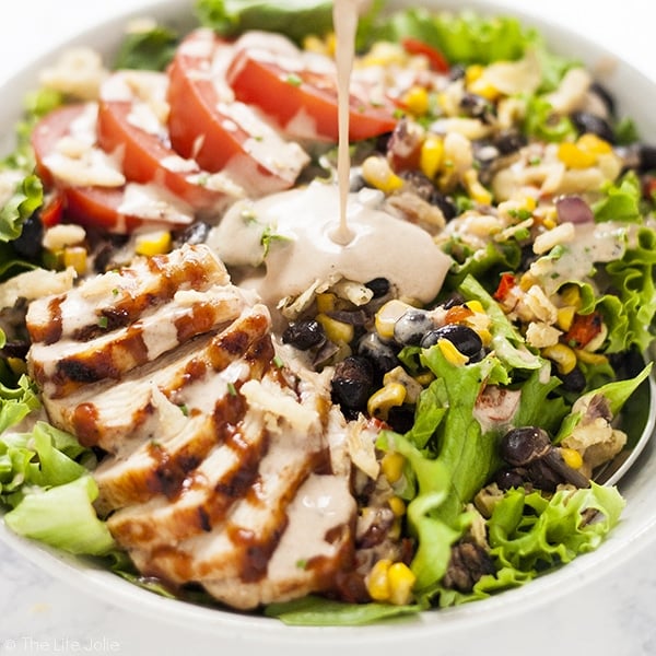 A square image of a close up image of bbq ranch dressing bring poured on a green salad with chicken, corn, black beans and tomatoes in a white bowl.
