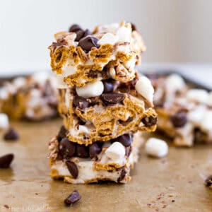 A square straight-on image of 3 s'mores bars stacked on top of each other with other bars behind them and chocolate chips and marshmallows.