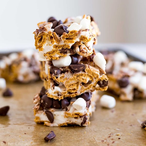 A square straight-on image of 3 s'mores bars stacked on top of each other with other bars behind them and chocolate chips and marshmallows.