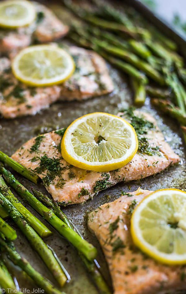 Three pieces of Lemon Dill Salmon lined up on the diagonal on a sheet pan with Asparagus on either side of it, the center piece of salmon is in focus and everything surrounding it is blurry. Each piece of fish has a lemon round on it.