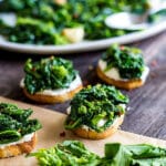 A square image of Broccoli Rabe and Ricotta Crostini with a yellow broccoli rabe blossom in the foreground and some other crostini and a plate of broccoli rabe in the background.