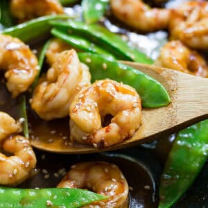 A square image of Shrimp and Snow Peas Stir Fry in a pan with a piece of shrimp on a wooden spoon.