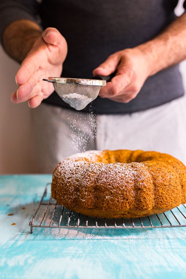 A man using a sieve to sprinkle powdered sugar onto a Poppy Seed Cake.