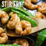 An image of Shrimp and Snow Peas Stir Fry in a pan with a piece of shrimp on a wooden spoon.