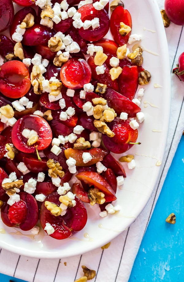 An over head image of the bottom of a plate of plums with goat cheese and walnuts on a white cloth and blue background.