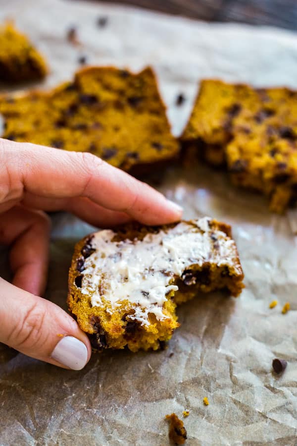 A hand holding a piece of Pumpkin Chocolate Chip Bread with butter on it and a bite take out of it.