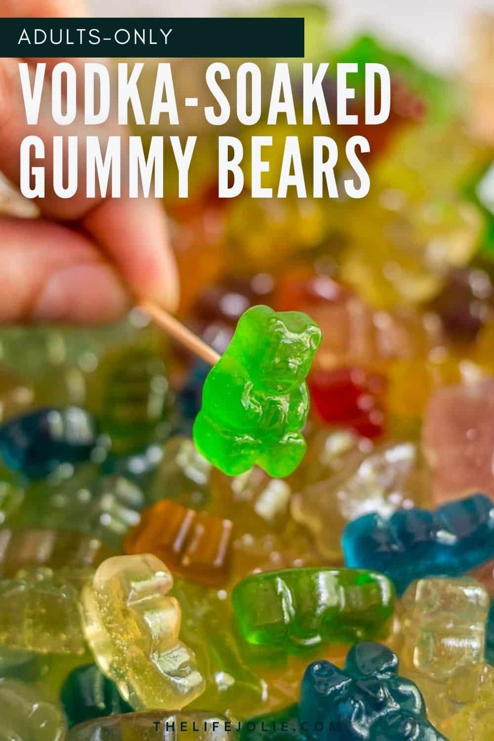 A hand holding a toothpick with a boozy gummy bear on it with a bunch of the gummy bears behind it.