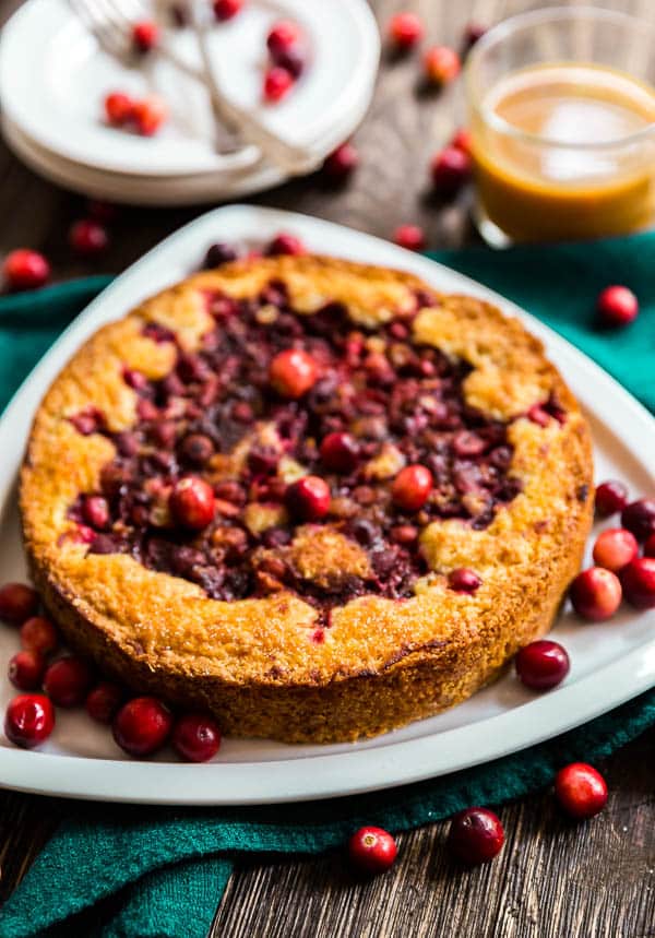 Orange Cranberry Torte with cranberries around it and a cup of coffee and some dessert plates in the background.