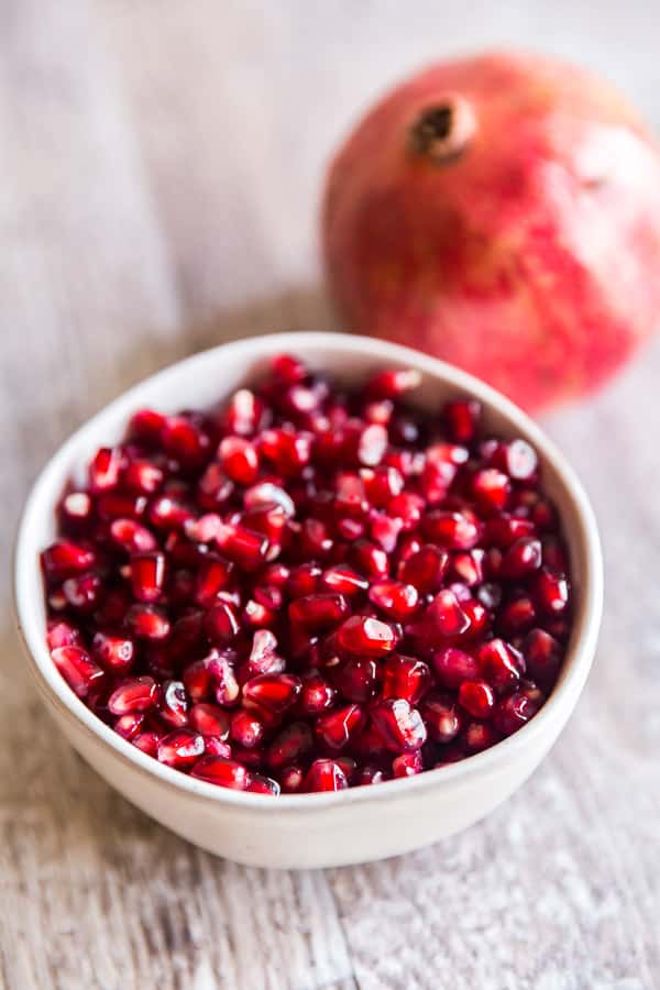 A bowl of pomegranate seeds with a whole fruit behind it for How to peel a pomegranate.