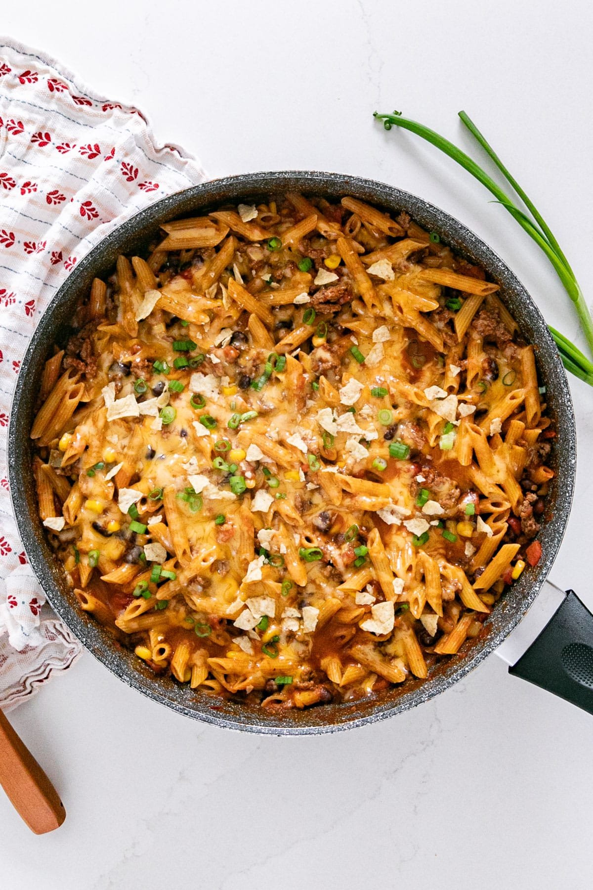 An overhead image of taco pasta in a skillet topped with crushed tortilla chips and chopped scallions.