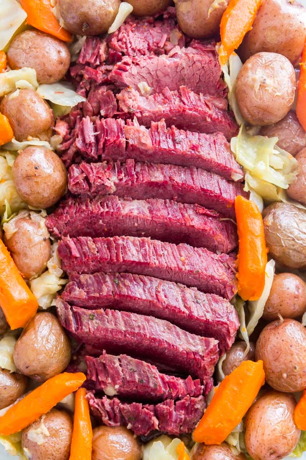 An overhead shot of sliced corned beef lined up on a plate surrounded by potatoes, carrots, and onions.