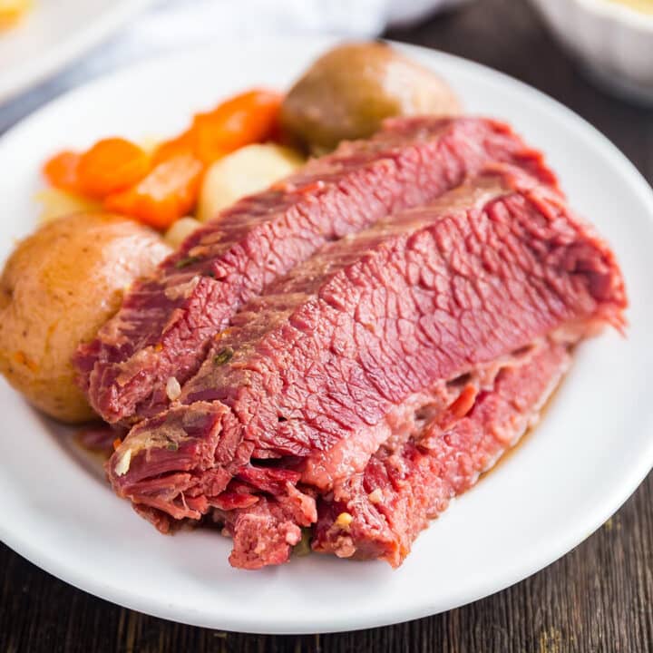 A square image of slices of corned beef on a white plate with potatoes and carrots in the background on the plate.