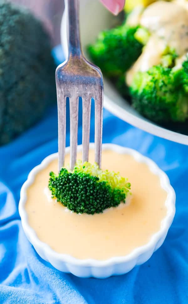 A Fork dipping broccol into a white ramekin of homemade cheese sauce with broccoli behind it.