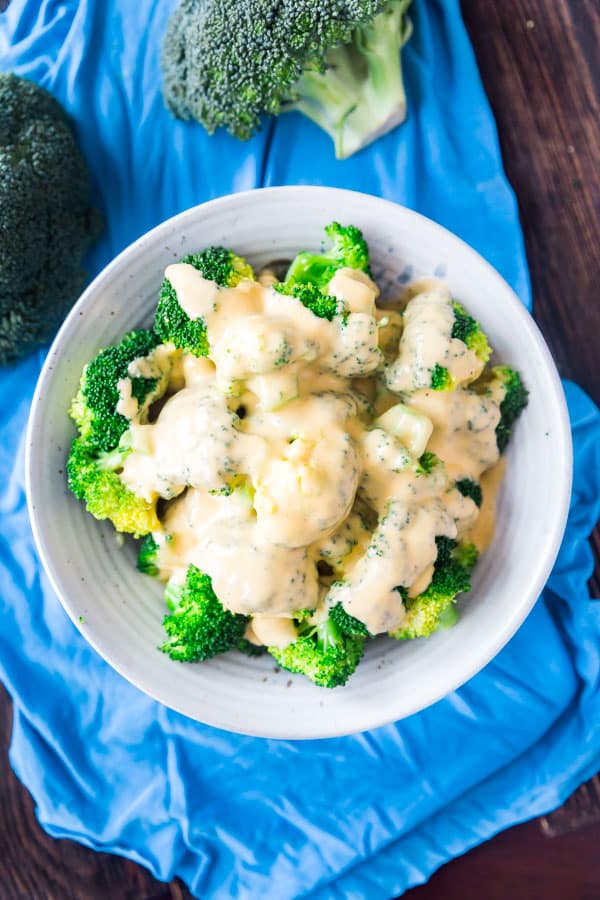 An overhead image of cheese sauce on steamed broccoli in a grey bowl on a blue towel with a broccoli floret above it.