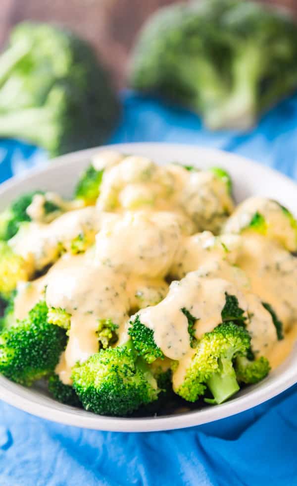 A bowl of broccoli with cheese sauce on a blue towel with broccoli florets in the background.