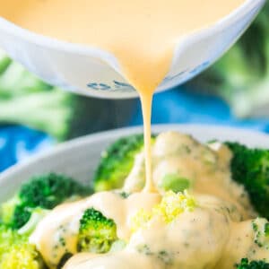 A square image of a white pot pouring cheese sauce over a bowl of broccoli with a blue towel behind it.