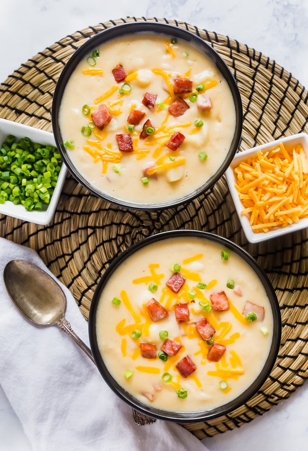 An overhead image of this Potato soup recipe in two bowls topped with ham on a rattan place mat with bowls of green onion and cheese nearby and a spoon on the side.