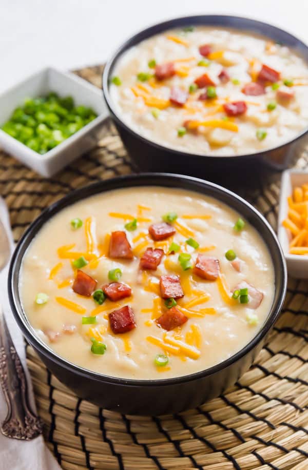 A side angle image of easy potato soup topped with ham, cheese and green onion with a bowl in focus up front and a second bowl behind it with a small bowl of green onions and shredded cheese in the background as well.