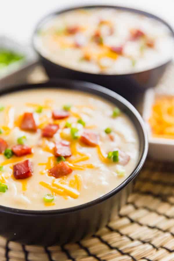 A close up of the side of a bowl of cheesy potato soup recipe with another bowl behind it and small bowls of green onion and shredded cheese on either side of it.