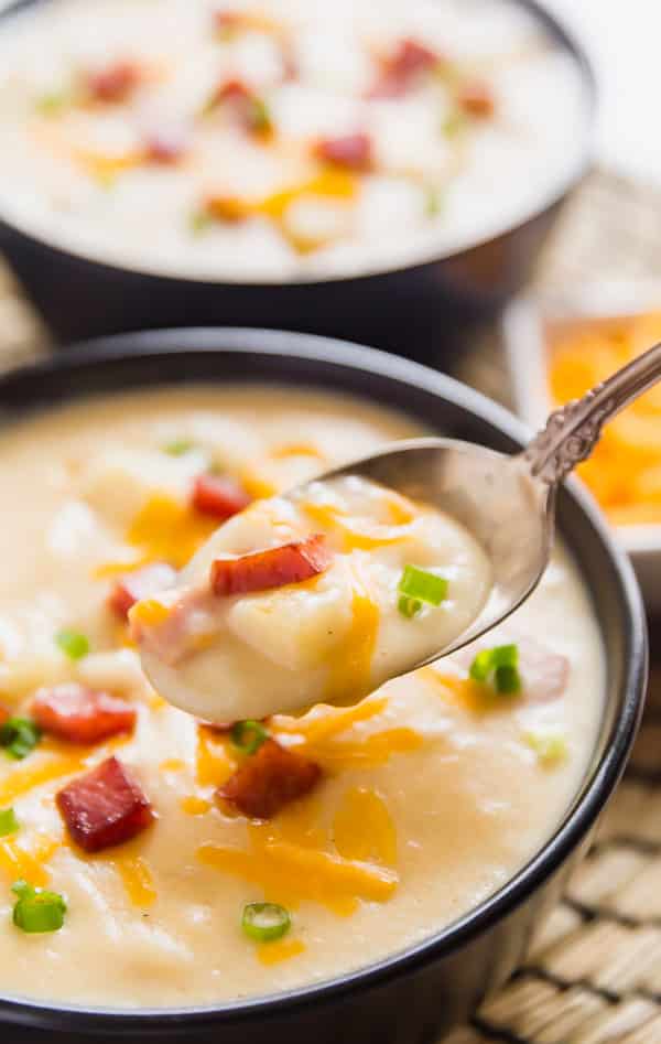 A close up image of a spoon holding a scoop of creamy potato soup with bowls of it in the background.
