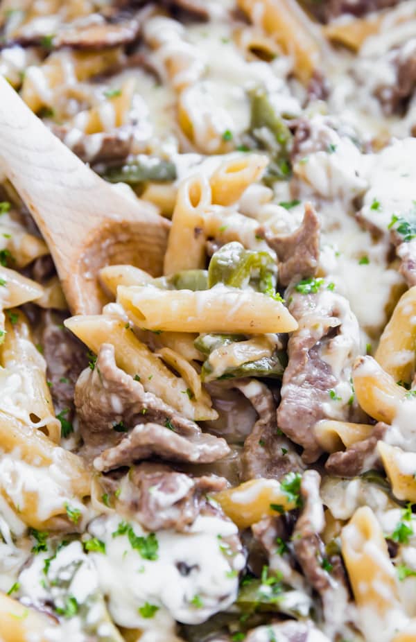 A close up image of a wooden spoon in a pan of steak pasta with penne, cheese, steak and peppers.
