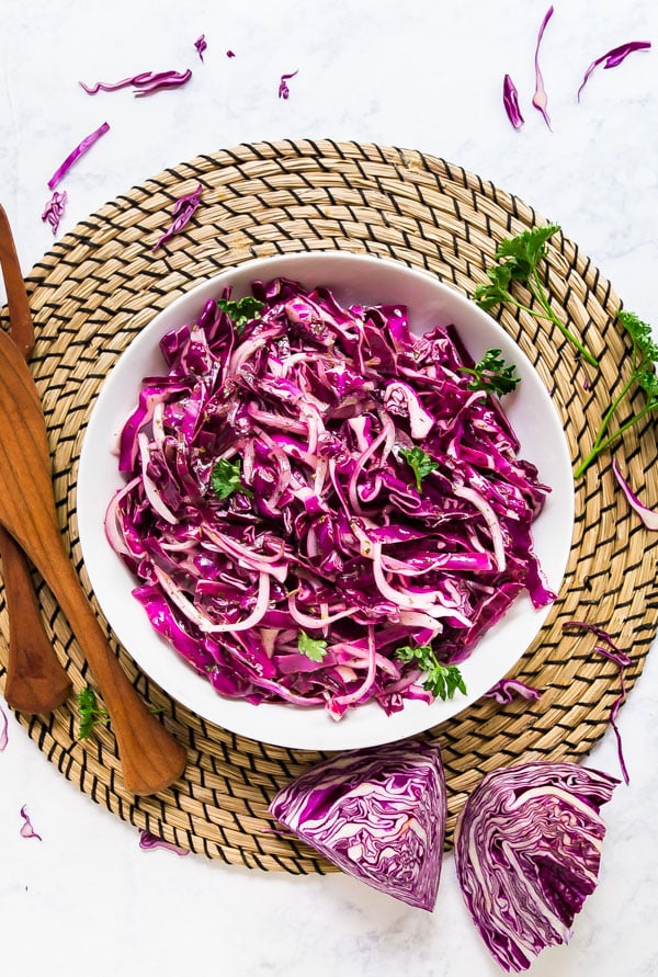 An overhead image of red cabbage salad in a white bowl on a woven placemat with shreds of red cabbage and a wooden spoon around it.