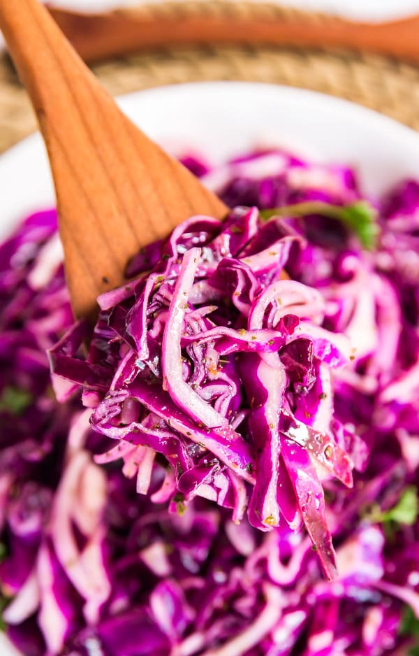 A wooden spoon holding up a scoop of red cabbage salad with the rest of the bowl in the background.