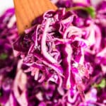 A square image of a wooden spoon holding up a scoop of red cabbage salad with the rest of the bowl in the background.