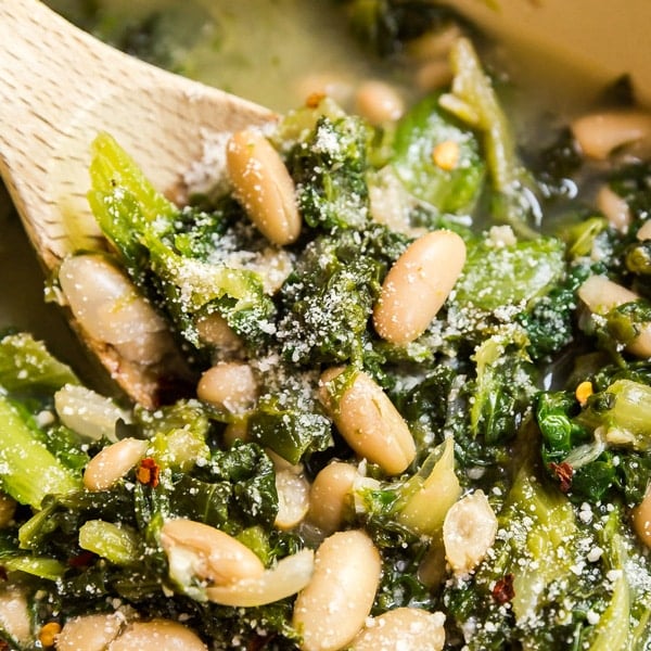 A square image of a wooden spoon in a pan of beans and greens.