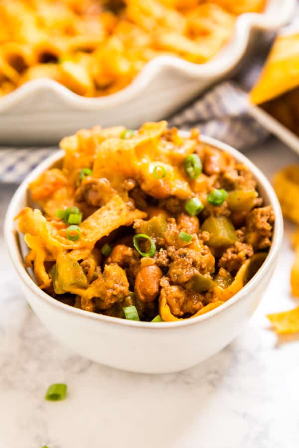A white bowl of Frito pie taco bake recipe with the pan of it in the background.