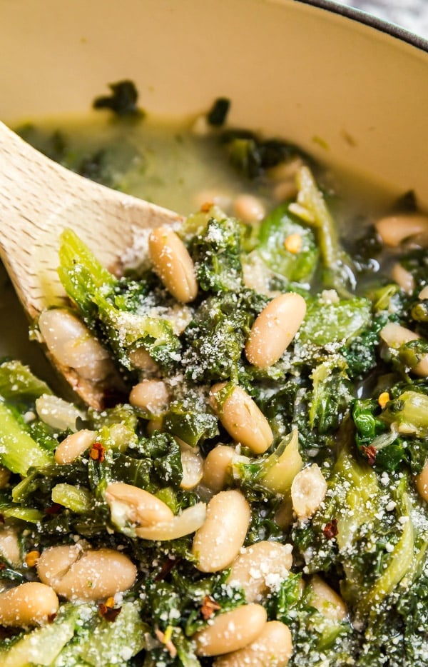 A close up image of beans and greens in a pan with a wooden spoon holding them up.