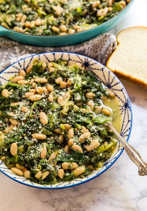 A bowl of escarole and beans with a spoon in it and the pan of it in the background. Also with bread on the side.