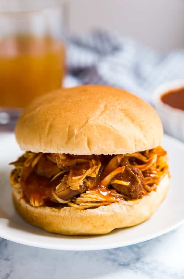 Bbq pulled chicken slow cooker style on a bun. on a white plate with bbq sauce, apple cider and a light blue cloth behind it out of focus.