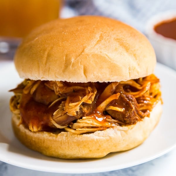 A square image of Bbq pulled chicken slow cooker style on a bun. on a white plate with bbq sauce, apple cider and a light blue cloth behind it out of focus.