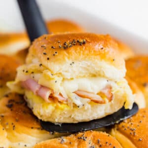 A square image of a ham and cheese slider on a spatula on top of a pan of other sliders.