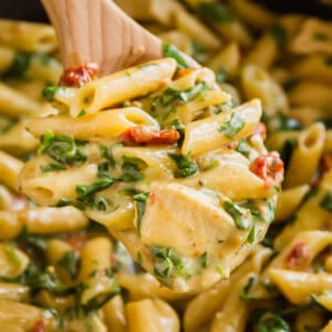A square image of a wooden spoon holding a scoop of florentine pasta with chicken with the rest of the pasta in the pan in the background.