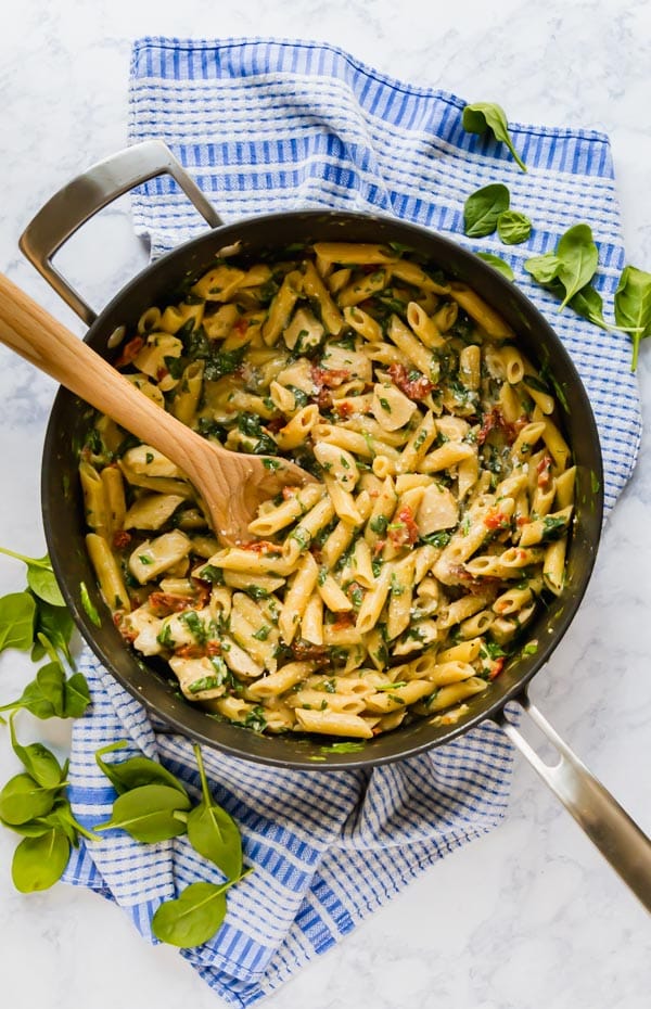 An overhead image of a pan of chicken pasta witha wooden spoon in it n a blue and white towel with fresh spinach around it.
