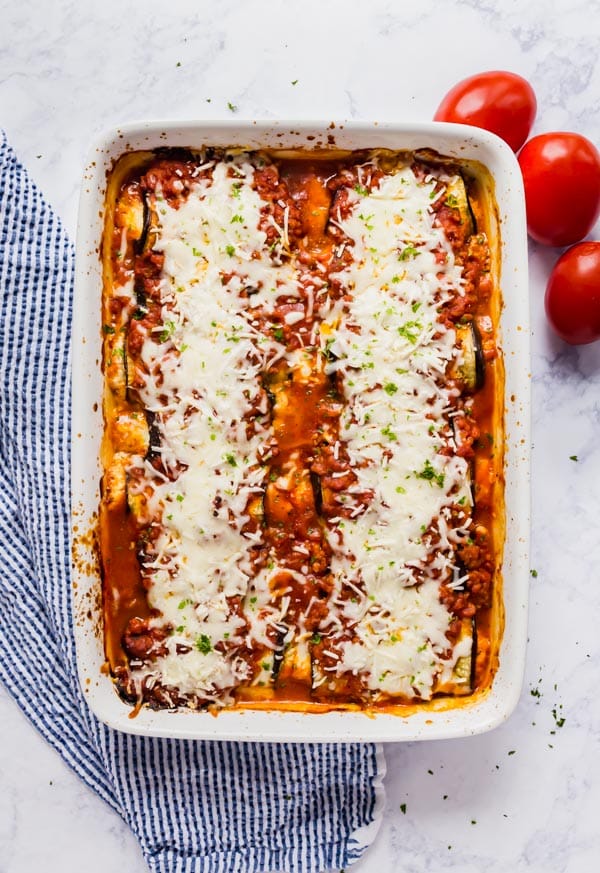 An overhead image of a plan of this eggplant rollatini recipe with a blue and white striped napkin next to it and tomatoes.
