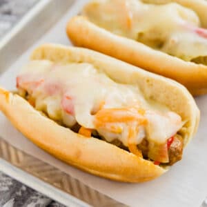 A square image of Buns with sausage and peppers and melted cheese on top on white rectangular plate.