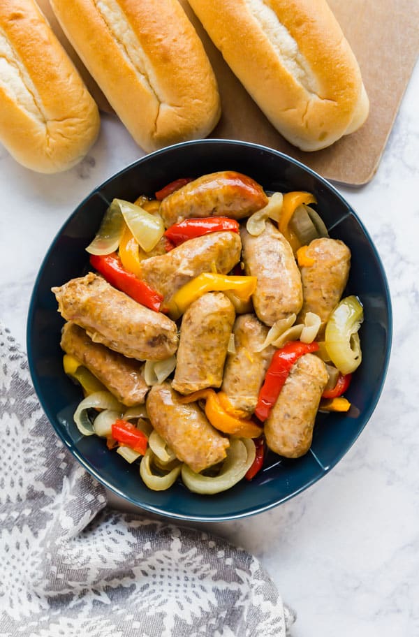 An overhead image of a blue bowl of sausage, peppers and onion with bun lined up next to it on a cutting board.