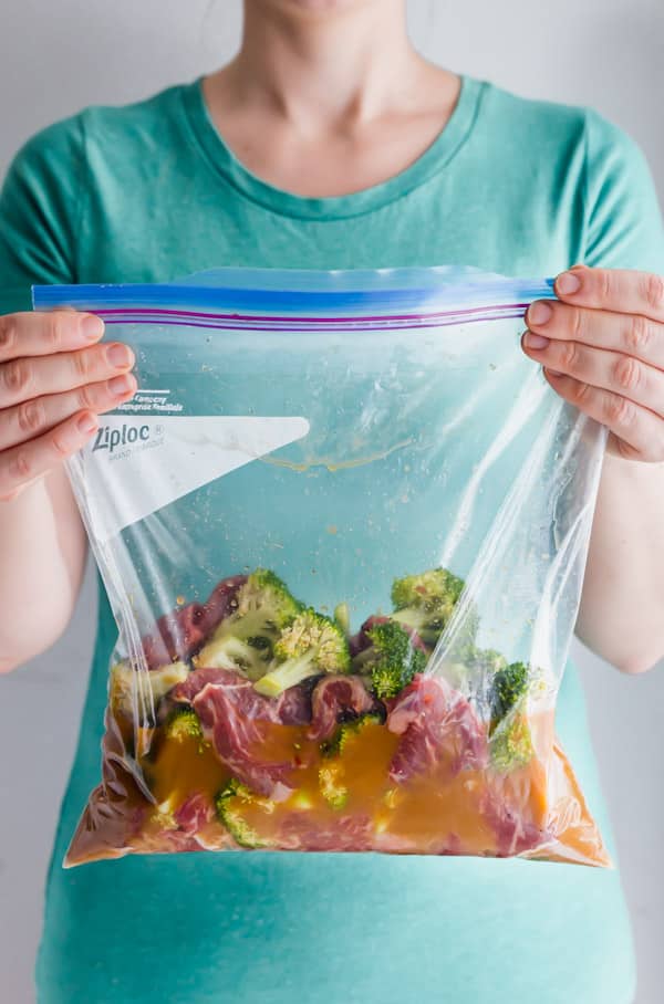 A straight-on image of a woman in a green shirt holding a zipper bag full of the raw beef and broccoli in the marinade.