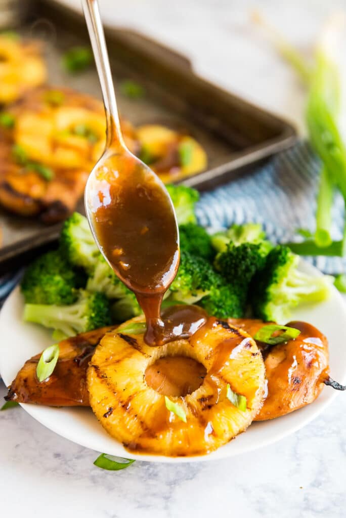 A side angle image of hawaiian chicken and pineapple on a plate with sauce being drizzled on top with a spoon.