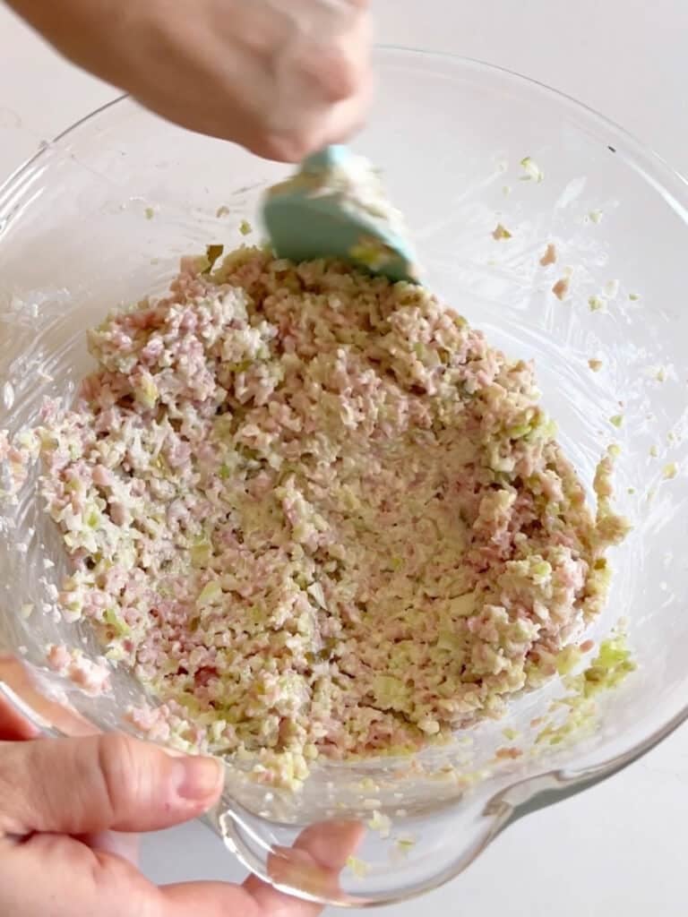 A hand mixing all the ham salad ingredients in a mixing bowl.