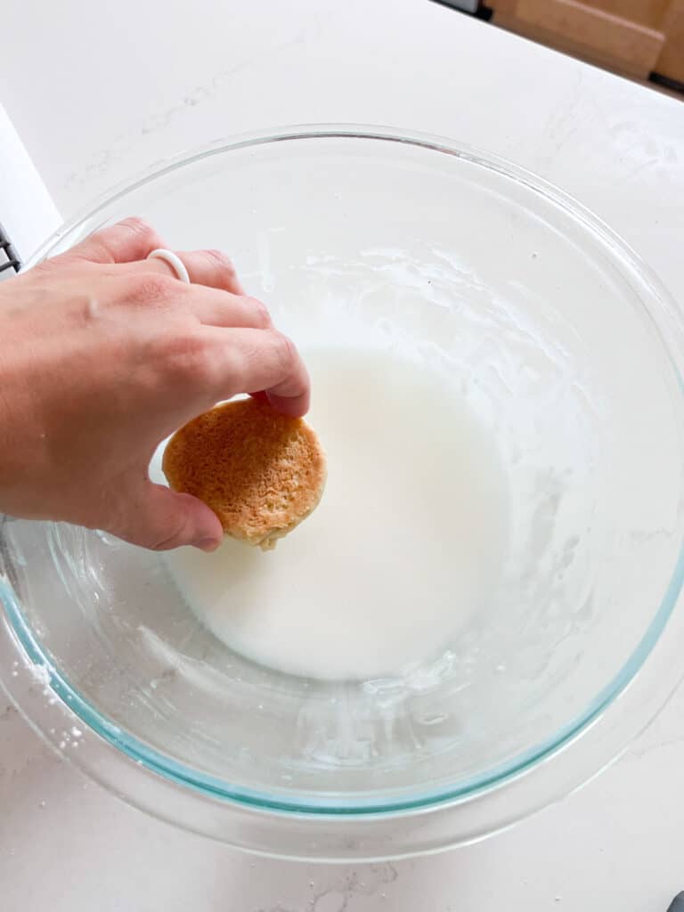 A hand dipping the top of a cookie into a bowl of glaze.