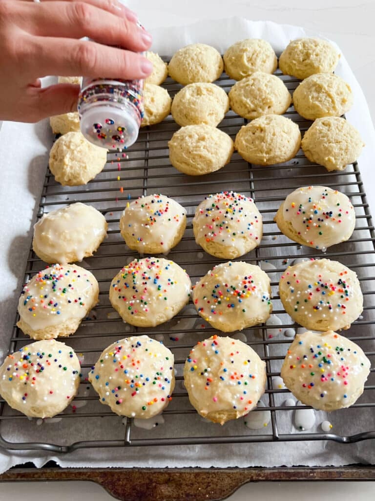 Cookies lined up on a cooling rack while a hand shakes sprinkles onto the glazed cookies.