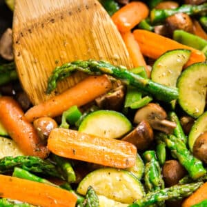 A square image of a close up inside of a pan of a bowl of vegetable medley with a wooden spoon in it.