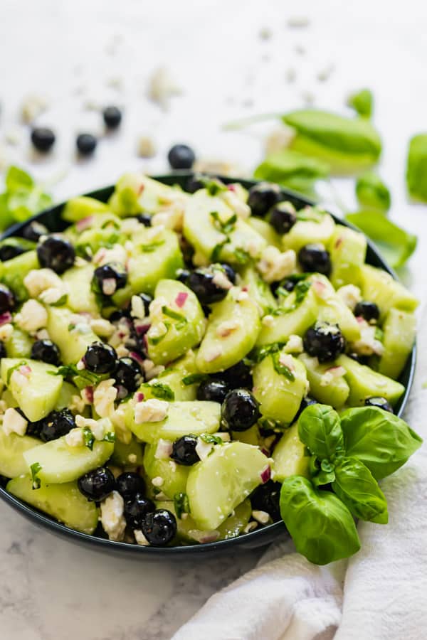 A bowl of cucumber salad with feta, blueberries and basil around it.