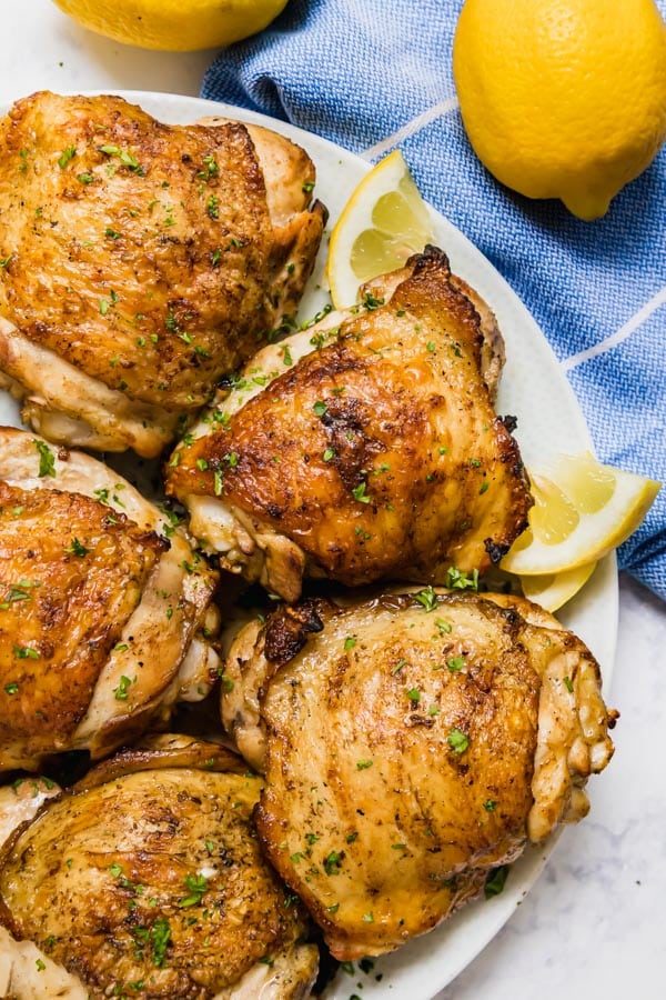 An overhead image of grilled chicken thighs on a plate with lemons next to it.
