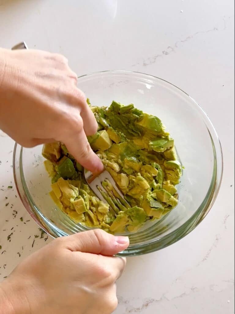 A hand mashing up avocado in a bowl with a fork.